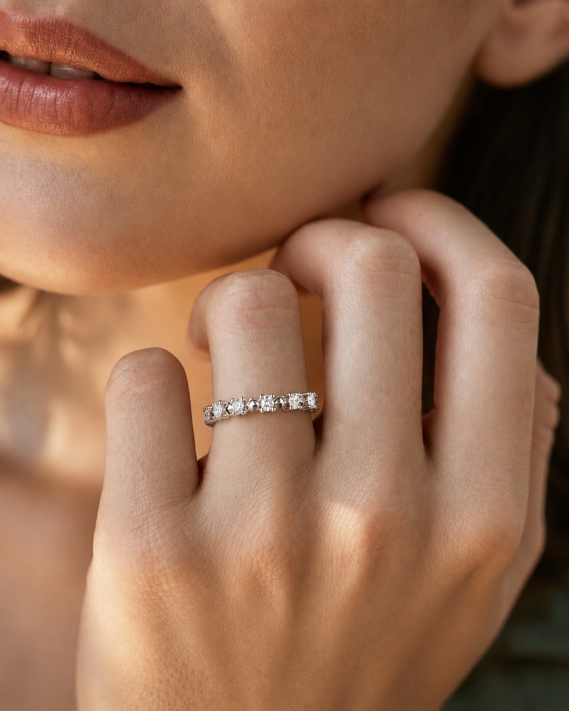 color_925_SIL,color_935_ARG,color_10K_WG, color_14K_WG &&A close-up lifestyle shot of a woman's hand wearing a delicate silver band ring with small sparkling stones, set against her cheek and jawline.