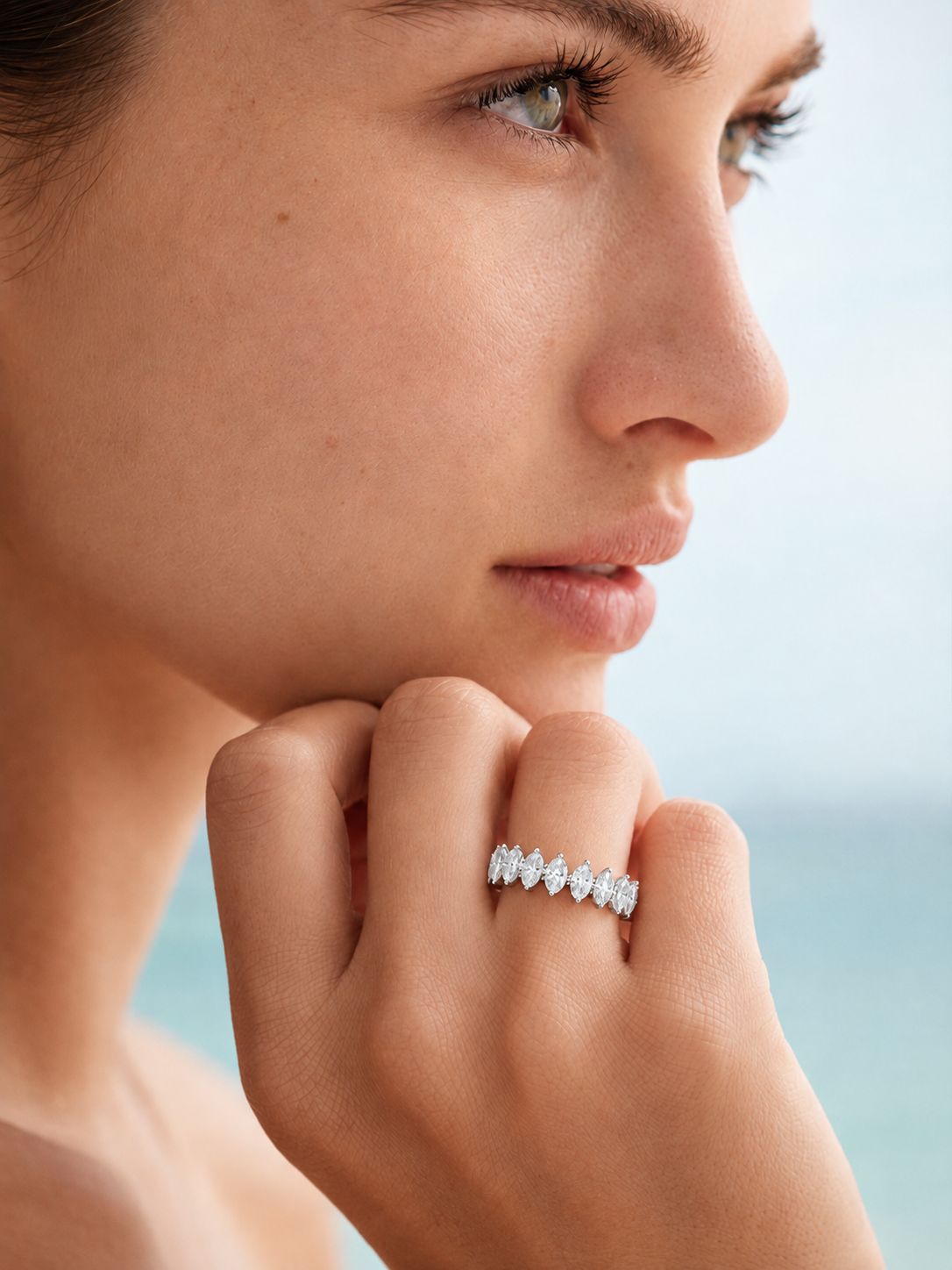 color_925_SIL,color_935_ARG,color_10K_WG, color_14K_WG &&A close-up lifestyle shot of a woman wearing a delicate silver band ring with a row of sparkling marquise-cut diamonds, set against a soft, blurred background of sky and sea.