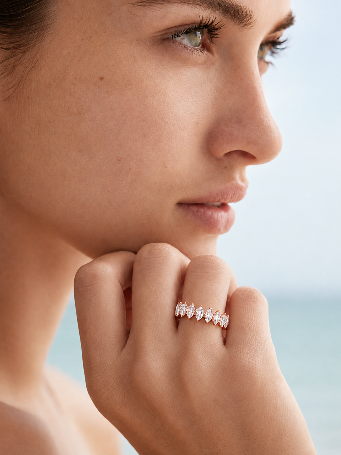 color_10K_RG, color_14K_RG &&A close-up lifestyle shot of a woman wearing a delicate rose gold band ring with a row of small clear marquise-cut gemstones. The background is a soft, out-of-focus coastal scene.