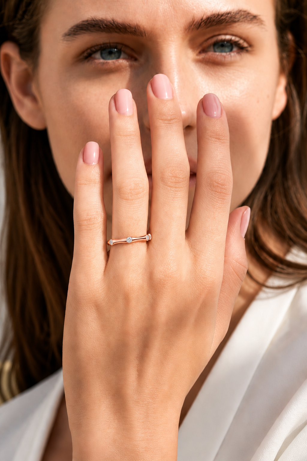 color_10K_RG, color_14K_RG &&A close-up lifestyle shot of a woman wearing a delicate rose gold ring with a small clear gemstone on her finger, with her hand covering part of her face.
