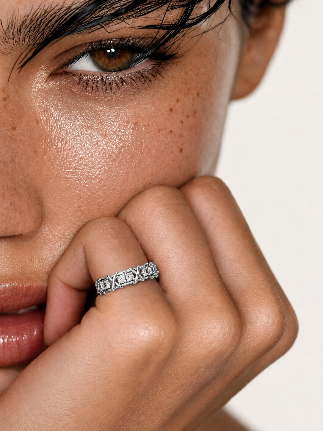 color_925_SIL,color_935_ARG,color_10K_WG, color_14K_WG &&A close-up lifestyle shot of a model wearing a silver band ring with a central clear gemstone and surrounding small clear stones, resting her chin on her hand against a clean white background.