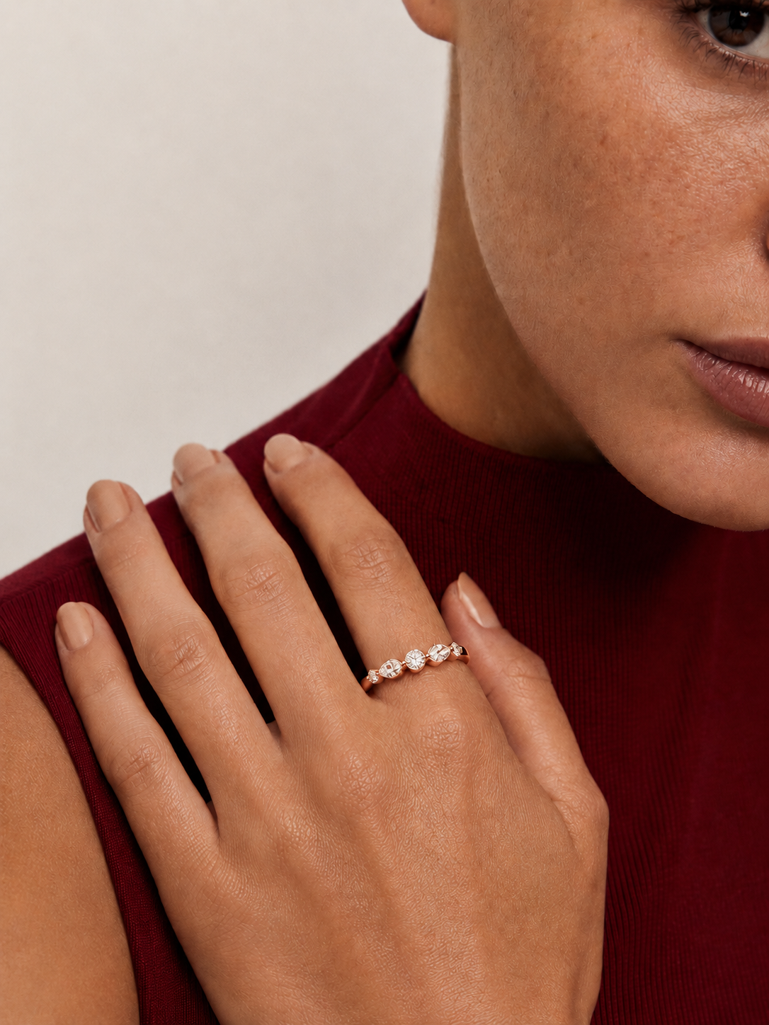 color_10K_RG, color_14K_RG &&A close-up lifestyle shot of a model's hand wearing a delicate rose gold ring with three small clear gemstones, set against a neutral light background and a dark red ribbed top.