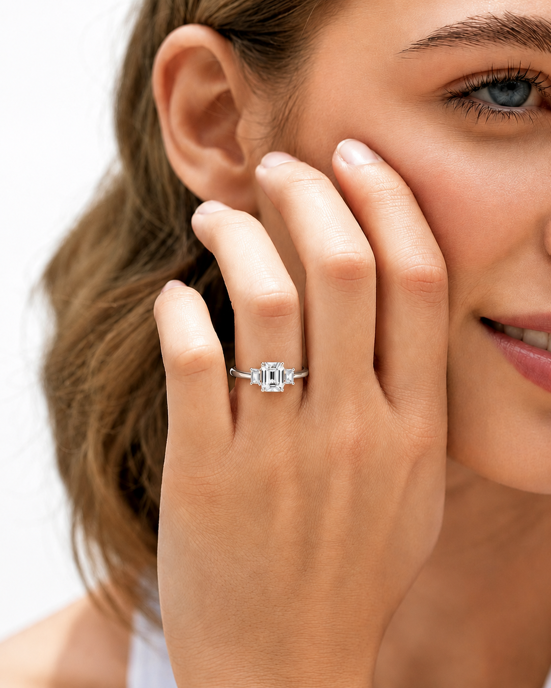 color_925_SIL,color_935_ARG,color_10K_WG, color_14K_WG &&A close-up lifestyle shot of a woman wearing a silver ring with a rectangular baguette-cut clear gemstone on her finger, with her hand resting against her cheek.
