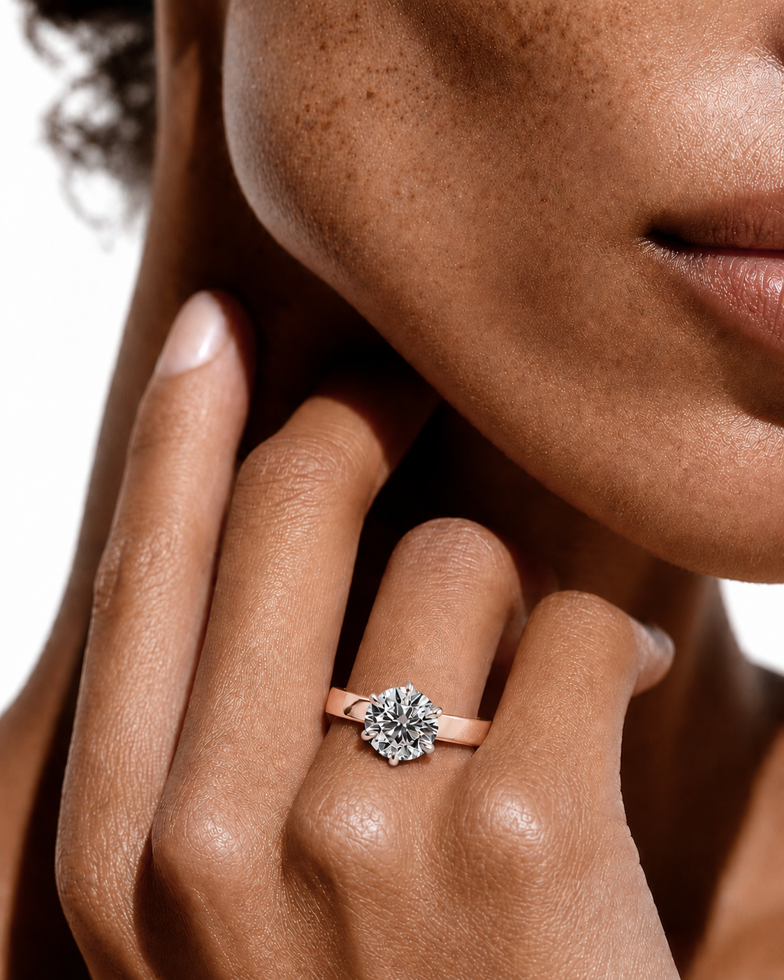 color_10K_RG, color_14K_RG &&A close-up lifestyle shot of a model's hand wearing a rose gold ring with a round-cut clear gemstone cluster, set against her cheek and neck.