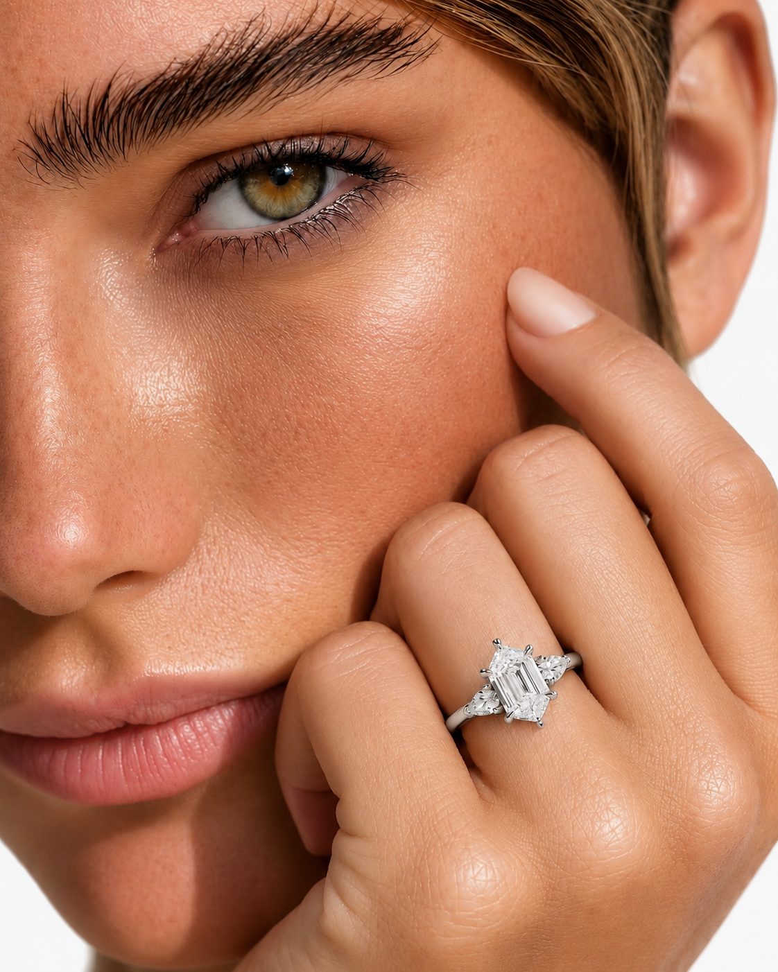 color_925_SIL,color_935_ARG,color_10K_WG, color_14K_WG &&A close-up lifestyle shot of a model wearing a silver emerald-cut ring on her finger, with her hand resting against her cheek against a clean white background.