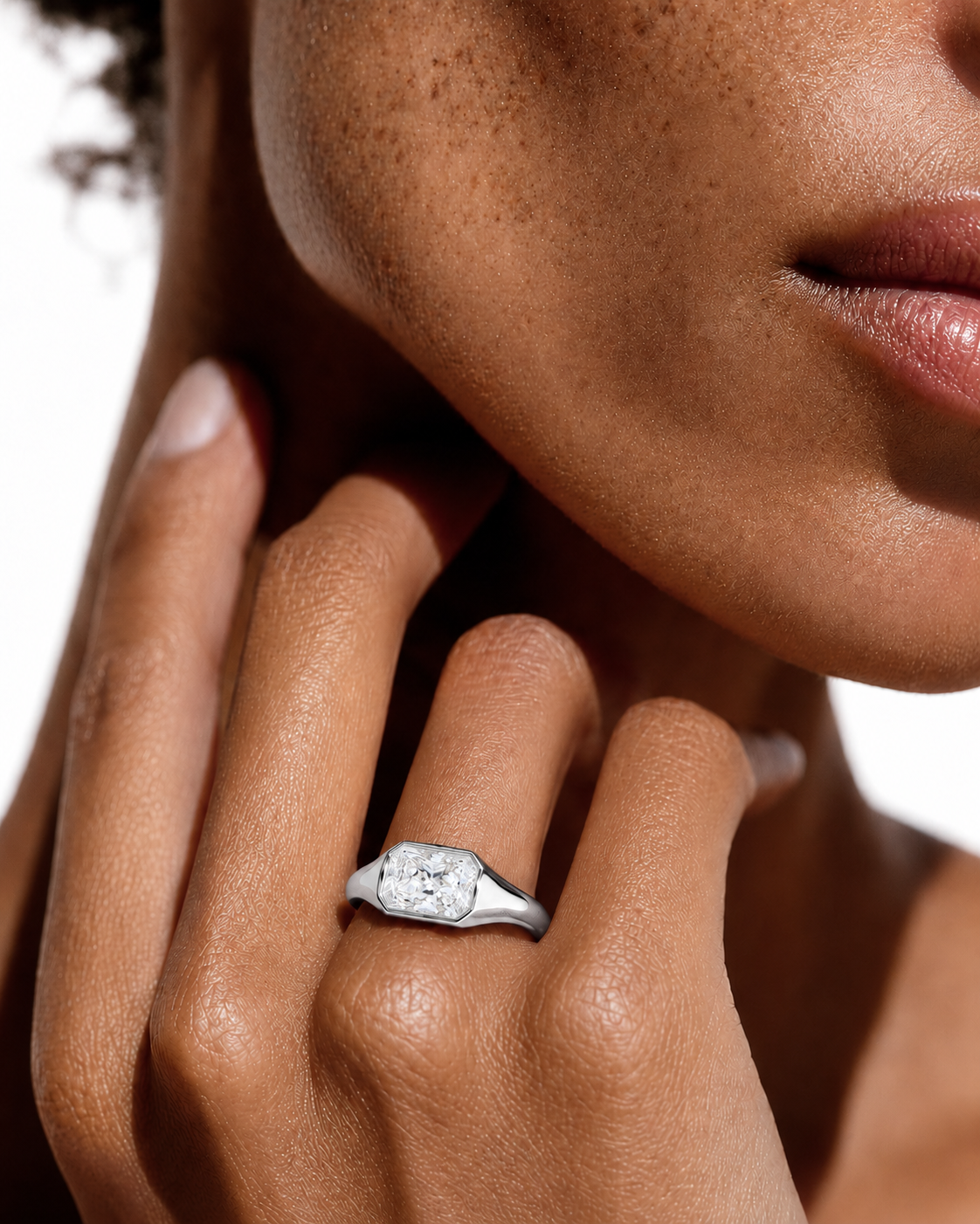 color_925_SIL,color_935_ARG,color_10K_WG, color_14K_WG &&A close-up lifestyle shot of a model wearing a silver ring with a cushion-cut diamond. The ring is shown on her finger, with her hand resting near her neck against a bright white background.
