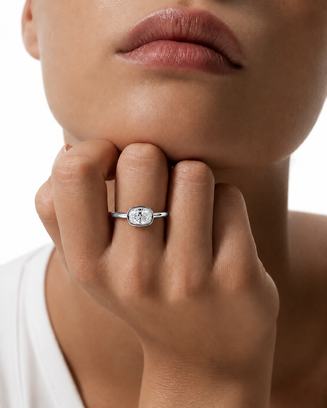 color_925_SIL,color_935_ARG,color_10K_WG, color_14K_WG &&A close-up lifestyle shot of a model wearing a silver band ring with a cushion-cut clear gemstone on her finger, with her hand resting on her chin against a white background.