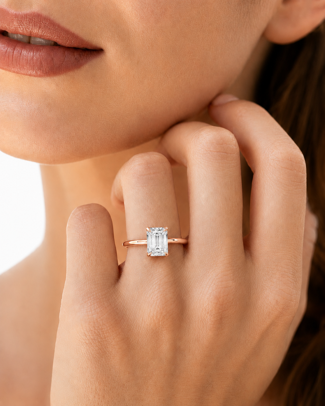 color_10K_RG, color_14K_RG &&A close-up lifestyle shot of a model wearing a rose gold engagement ring with a rectangular baguette-cut clear gemstone. The ring is shown on the model's finger, with her hand and lower face visible against a bright, clean background.