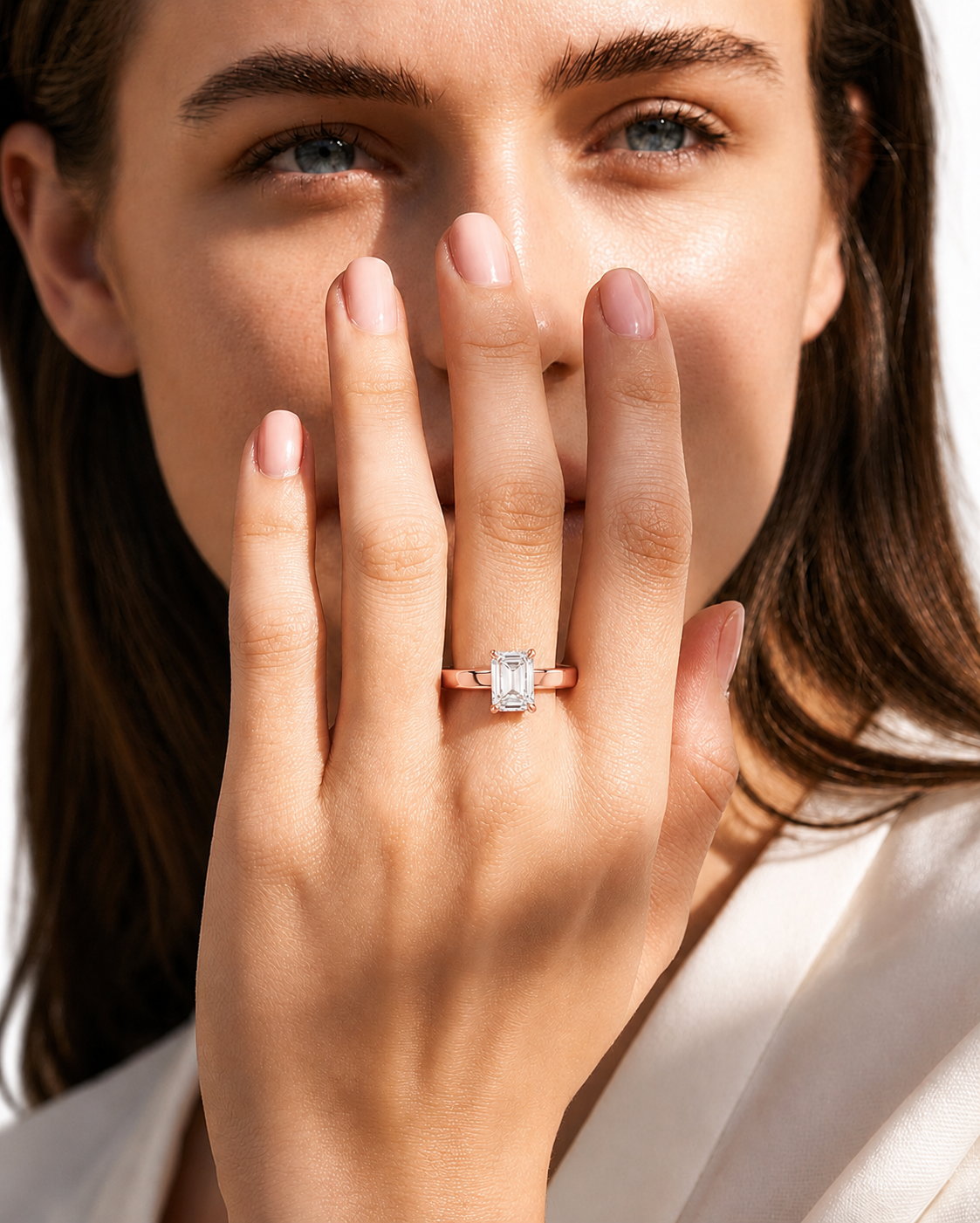 color_10K_RG, color_14K_RG &&A close-up lifestyle shot of a woman wearing a rose gold engagement ring with a rectangular emerald-cut diamond. The ring is shown on her finger, with her hand covering part of her face against a bright, clean white background.