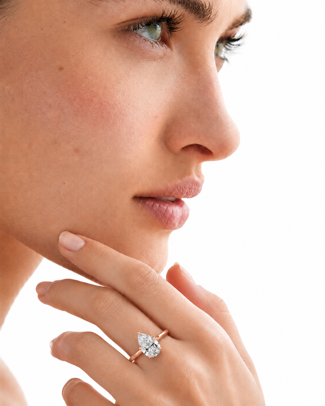 color_10K_RG, color_14K_RG &&A close-up side profile of a woman wearing a pear-cut diamond ring on a rose gold band, with her hand resting near her chin against a white background.