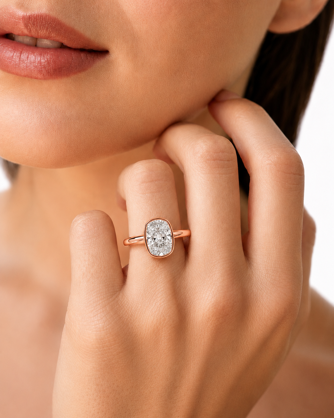 color_10K_RG, color_14K_RG &&A close-up lifestyle shot of a model wearing a rose gold ring with a cushion-cut diamond. The ring is shown on her finger, with her hand resting near her chin against a clean white background.