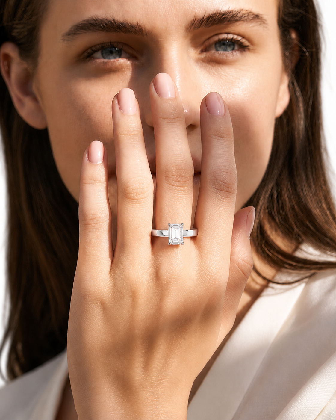 color_925_SIL,color_935_ARG,color_10K_WG, color_14K_WG &&A close-up lifestyle shot of a woman wearing a silver engagement ring with a rectangular emerald-cut center stone on her finger, with her hand covering part of her face.