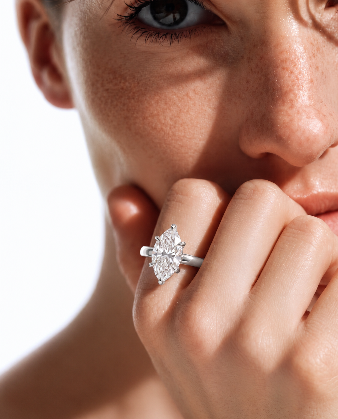 color_925_SIL,color_935_ARG,color_10K_WG, color_14K_WG &&A close-up lifestyle shot of a model's face and hand wearing a silver ring with a marquise-cut diamond. The ring is shown on the ring finger against a bright white background.