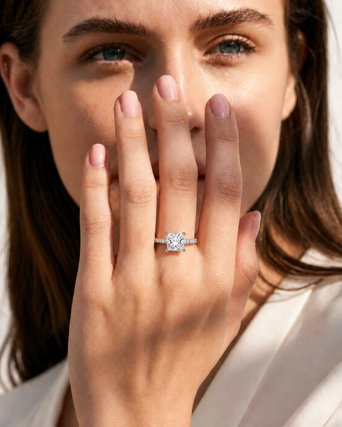 color_925_SIL,color_935_ARG,color_10K_WG, color_14K_WG &&A close-up lifestyle shot of a woman wearing a diamond engagement ring on her finger, holding her hand near her face. The ring features a round brilliant-cut center stone set in a silver-toned band with pavé-set side stones.