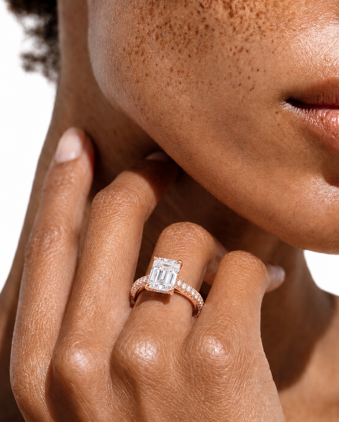 color_10K_RG, color_14K_RG &&A close-up lifestyle shot of a model's hand resting on her neck, wearing a rose gold ring with a rectangular emerald-cut diamond and a pavé band.