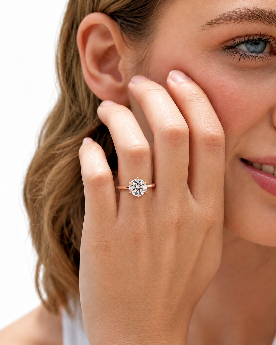 color_10K_RG, color_14K_RG &&A close-up lifestyle shot of a woman wearing a rose gold engagement ring with a round-cut clear gemstone. The ring is shown on her finger as she gently touches her cheek against a clean white background.