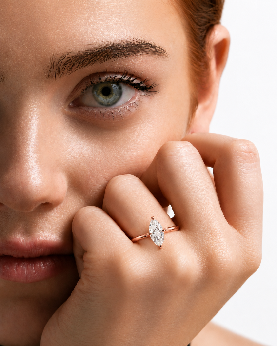 color_10K_RG, color_14K_RG &&A close-up lifestyle shot of a woman with red hair resting her hand near her face, wearing a rose gold ring with a marquise-cut clear gemstone on her finger.