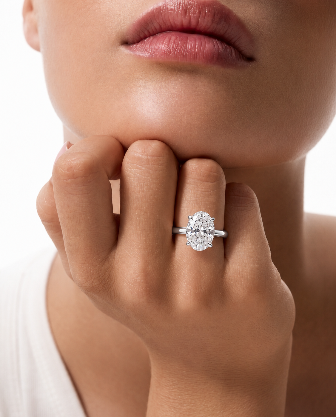 color_925_SIL,color_935_ARG,color_10K_WG, color_14K_WG &&A close-up lifestyle shot of a model wearing a silver engagement ring with a round-cut clear gemstone on her finger, with her hand resting on her chin against a white background.