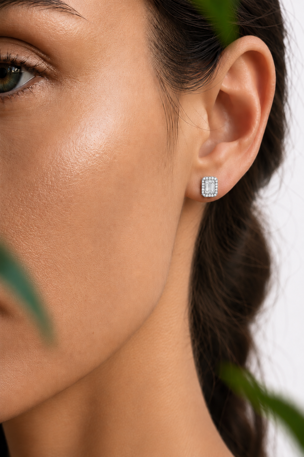 color_925_SIL,color_935_ARG,color_10K_WG, color_14K_WG &&A close-up of a woman's ear wearing a rectangular silver stud earring with a border of small clear stones, set against a light background with soft green leaves in the foreground.