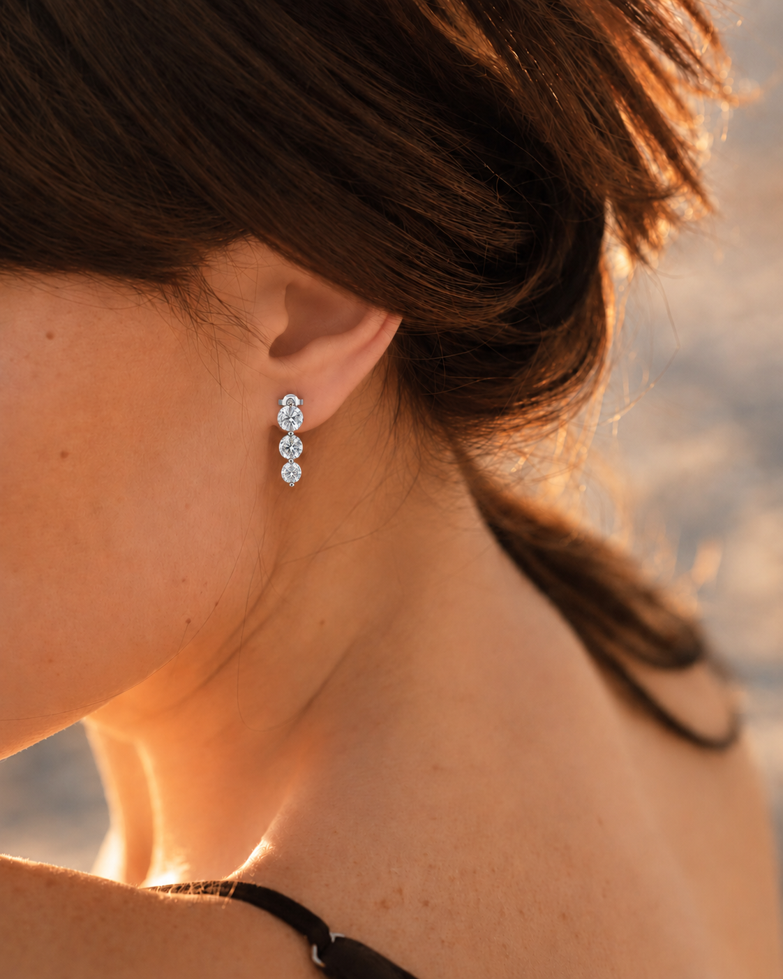 color_925_SIL,color_935_ARG,color_10K_WG, color_14K_WG &&A close-up lifestyle shot of a woman wearing a small silver-toned dangle earring with three clear gemstones, set against a warm, blurred outdoor background.